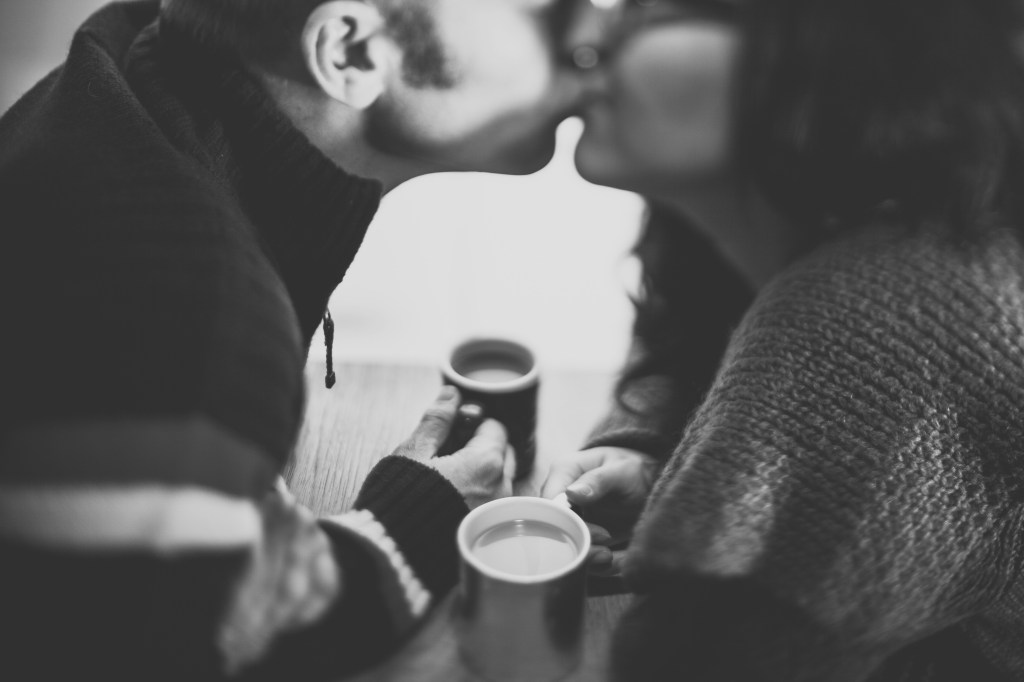 Greyscale photo of a couple kissing across a table. Their faces blur together. They're each holding a coffee mug.